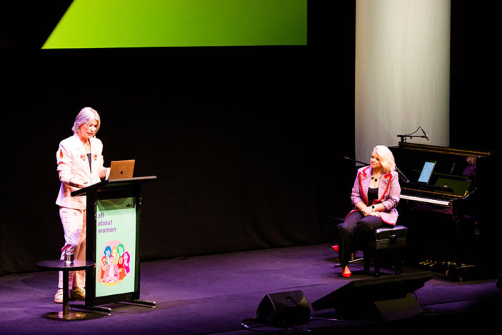 A woman dressed in a pink suit stands behind a podium while another woman watches while seated at a piano.