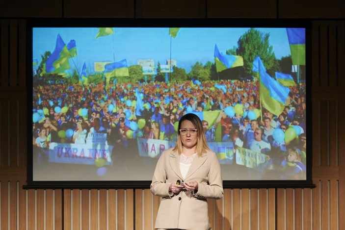 A woman stands in front of a presentation which shows crowds of people with banners.