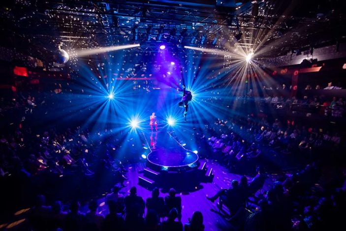 Wide shot of a dark room with spotlights shinning on a man suspended in mid-air by a rope.