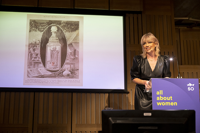 A woman stands behind a podium to the right of a screen which is showing an image of an old fashioned looking bottle of skincare. The woman is presenting to a crowd.