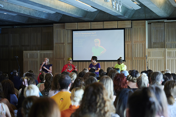 A crowd sits in a room listening to three women sat on a stage with a screen behind them.