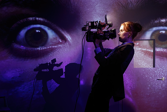 A woman holds a camera up to her face. Projected on the wall behind her is the close up view of her eyes.