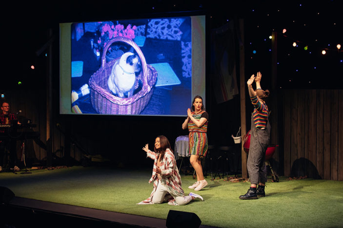 Three comedians perform on stage in front of a slide projection of a cat in a basket. 